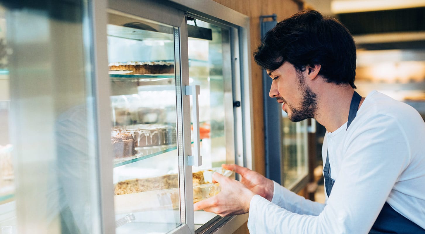 A supermarket clerk wearing an apron stocks the refrigerated section with a tray of cakes.